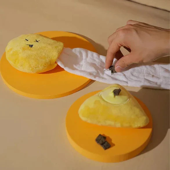 Hand interacting with yellow plush toy on orange base, connected by white fabric. Pet training aid, enrichment toy, interactive play.