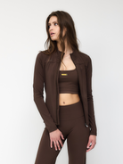 Woman in brown activewear set, featuring a zip-up jacket, sports bra, and leggings, posing against a white background. Fashion, fitness apparel.