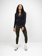 Woman wearing a black zip blouse and olive green leggings, standing against a white background. Fashionable activewear outfit with sneakers.