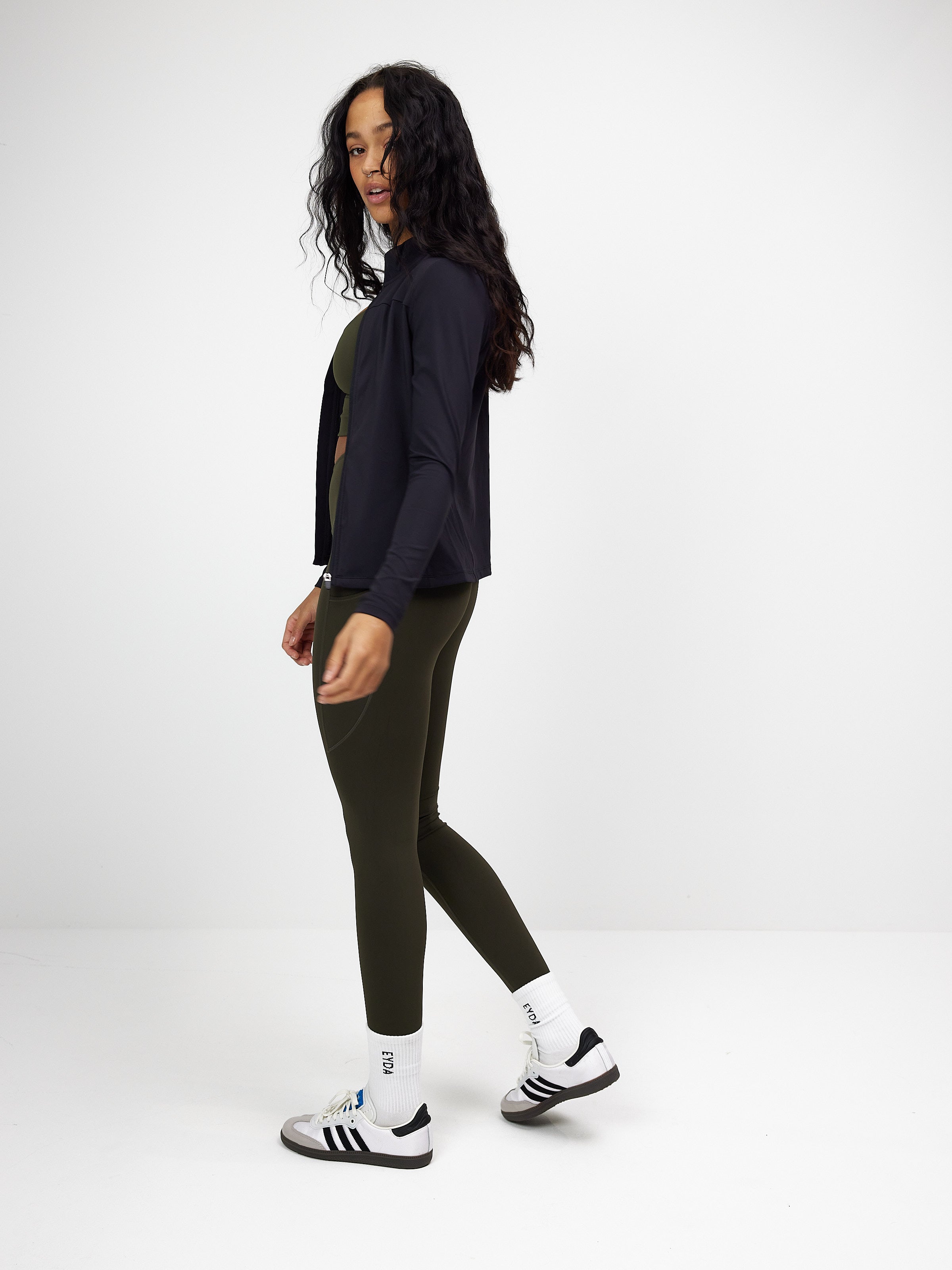 Woman wearing black zip blouse, olive green leggings, and white sneakers with black stripes, standing against a plain white background. Fashion, casual wear.