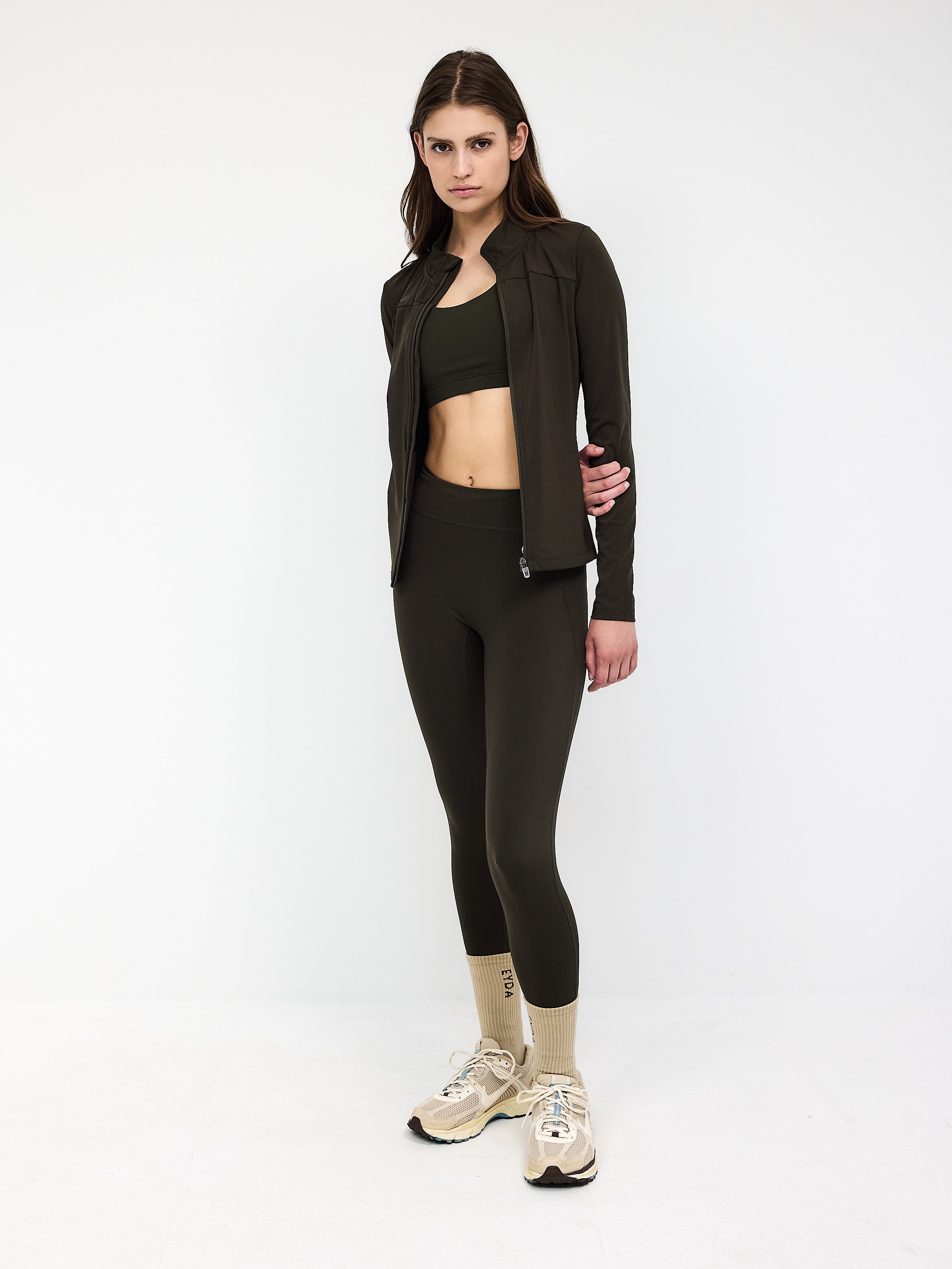 Woman in dark olive activewear set, featuring a zip-up jacket, sports bra, and leggings, paired with beige sneakers. Fitness fashion outfit.