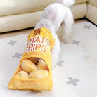 Dog with head inside potato chips bag toy on living room floor. Plush snack-themed pet toy, playful scene, white fluffy dog, home decor.