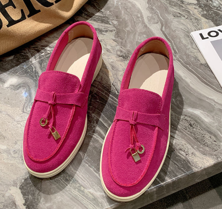 Pink suede loafers with tassel detail on a marble surface, next to a beige bag and a magazine. Stylish footwear, fashion accessories, casual shoes.