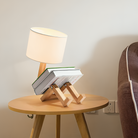 Modern wooden table lamp with a unique book-shaped design on a round wooden side table, next to a brown sofa. Ideal for stylish home decor.