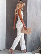 Blonde woman in elegant white lace jumpsuit holding beige clutch, standing against a modern gray wall. Fashionable summer outfit, stylish look.