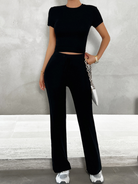 Woman wearing a stylish black two-piece outfit with a crop top and wide-leg pants, accessorized with a white handbag and sneakers. Fashionable casual wear.