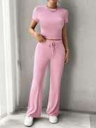 Woman wearing a pink two-piece lounge set with short-sleeve crop top and drawstring pants, paired with white sneakers, standing indoors.