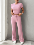 Woman wearing a pink two-piece lounge set with short-sleeve top and drawstring pants, accessorized with a necklace and watch, standing indoors.