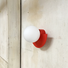 Modern red wall sconce with a round white globe light fixture, mounted on a textured beige wall. Contemporary lighting design, minimalist decor.