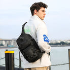 Man wearing a stylish black tactical sling backpack with multiple compartments, standing outdoors in a white jacket. Ideal for urban travel and adventure.