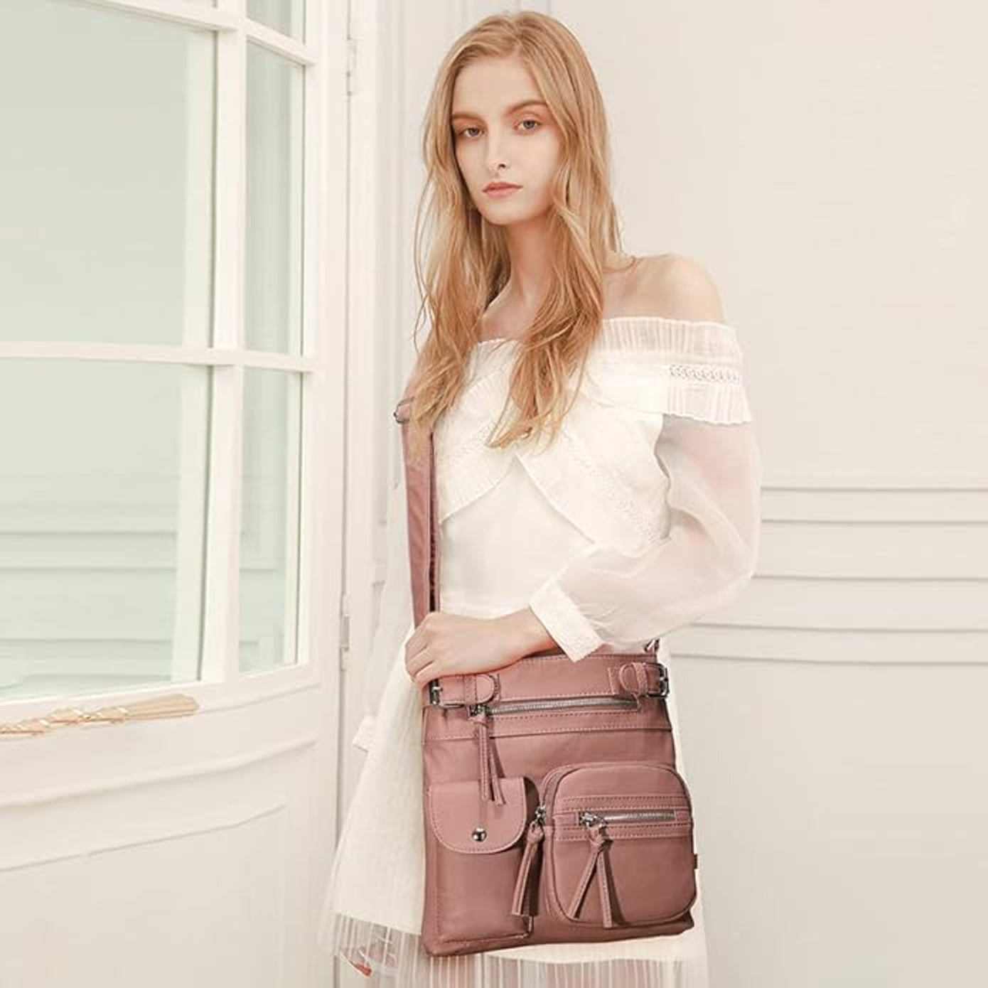 Woman in white dress holding a stylish pink crossbody bag with multiple pockets, standing indoors. Fashionable women's accessory, elegant design.