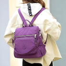 Purple backpack with multiple pockets and silver zippers, worn by a person in a white hoodie. Stylish, functional bag for travel or daily use.