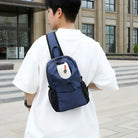 Man wearing a blue crossbody backpack with a white t-shirt, standing outside a modern building. Stylish urban fashion accessory for men.