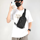 Man wearing a black crossbody sling bag, white graphic t-shirt, and black face mask, taking a selfie with a smartphone. Fashionable casual streetwear.