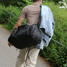 Man walking on a path carrying a black travel duffel bag and a denim jacket over his shoulder, surrounded by lush greenery.