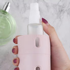 Hand holding a pink portable mist sprayer bottle on a marble surface, with a green perfume bottle nearby. Skincare, beauty, travel essentials.