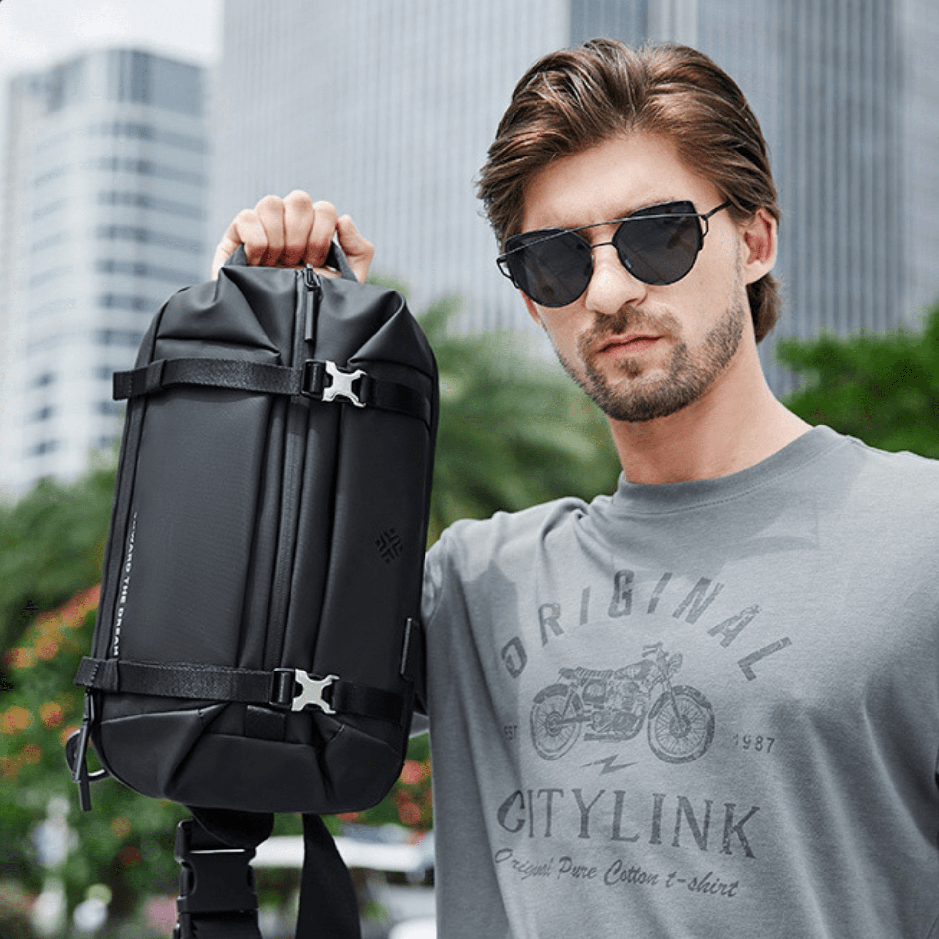 Man holding sleek black waterproof backpack in urban setting; stylish accessory for travel and commuting; modern design with secure buckles.