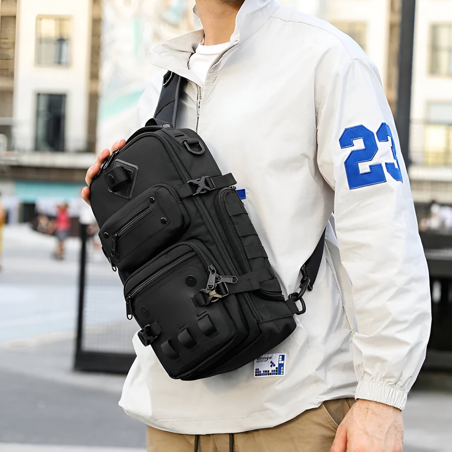 Man wearing a white jacket with blue "23" and carrying a black tactical sling bag. Urban street style, crossbody bag, outdoor fashion accessory.