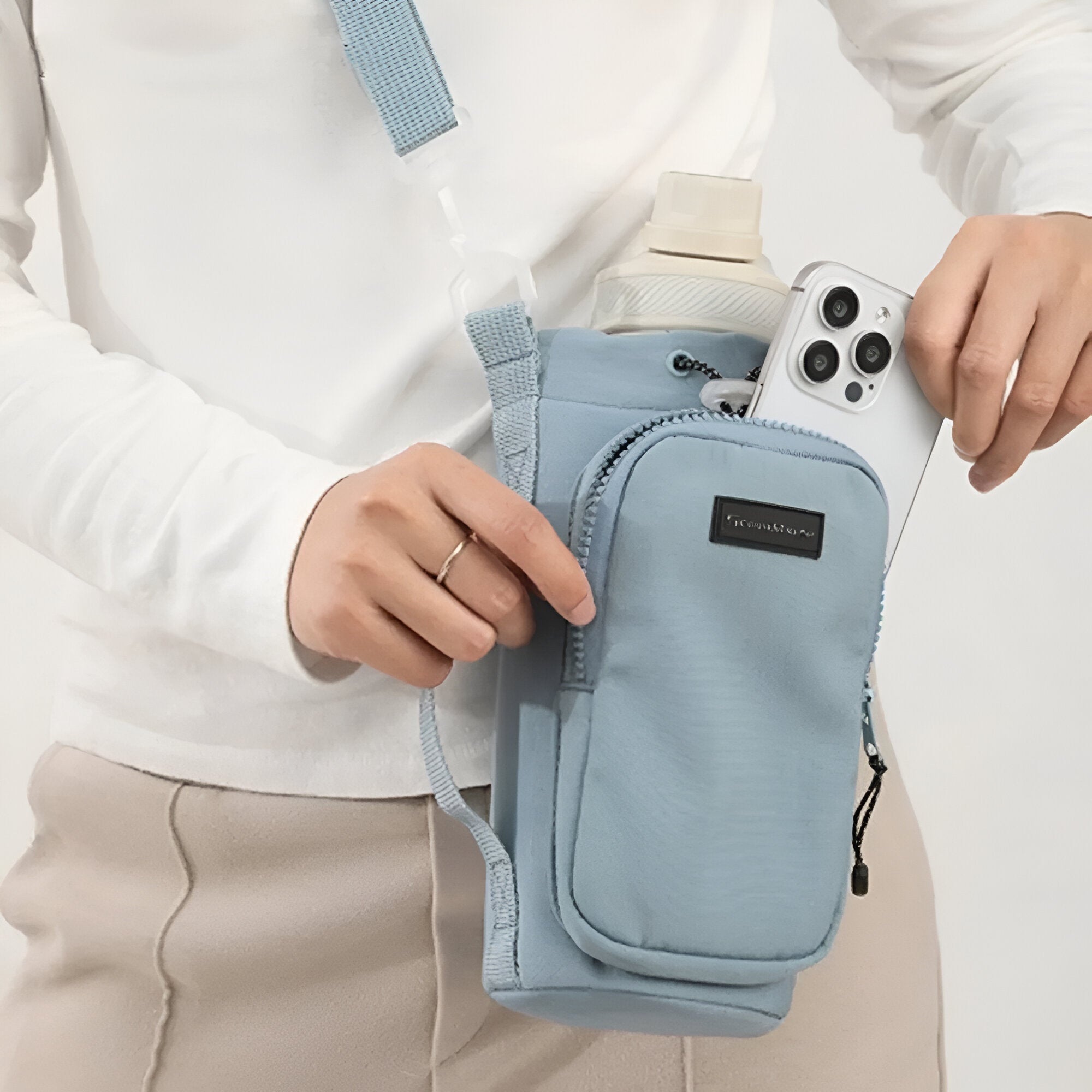 Person holding a light blue crossbody water bottle holder with a smartphone pocket, ideal for travel and outdoor activities. Stylish, functional accessory.