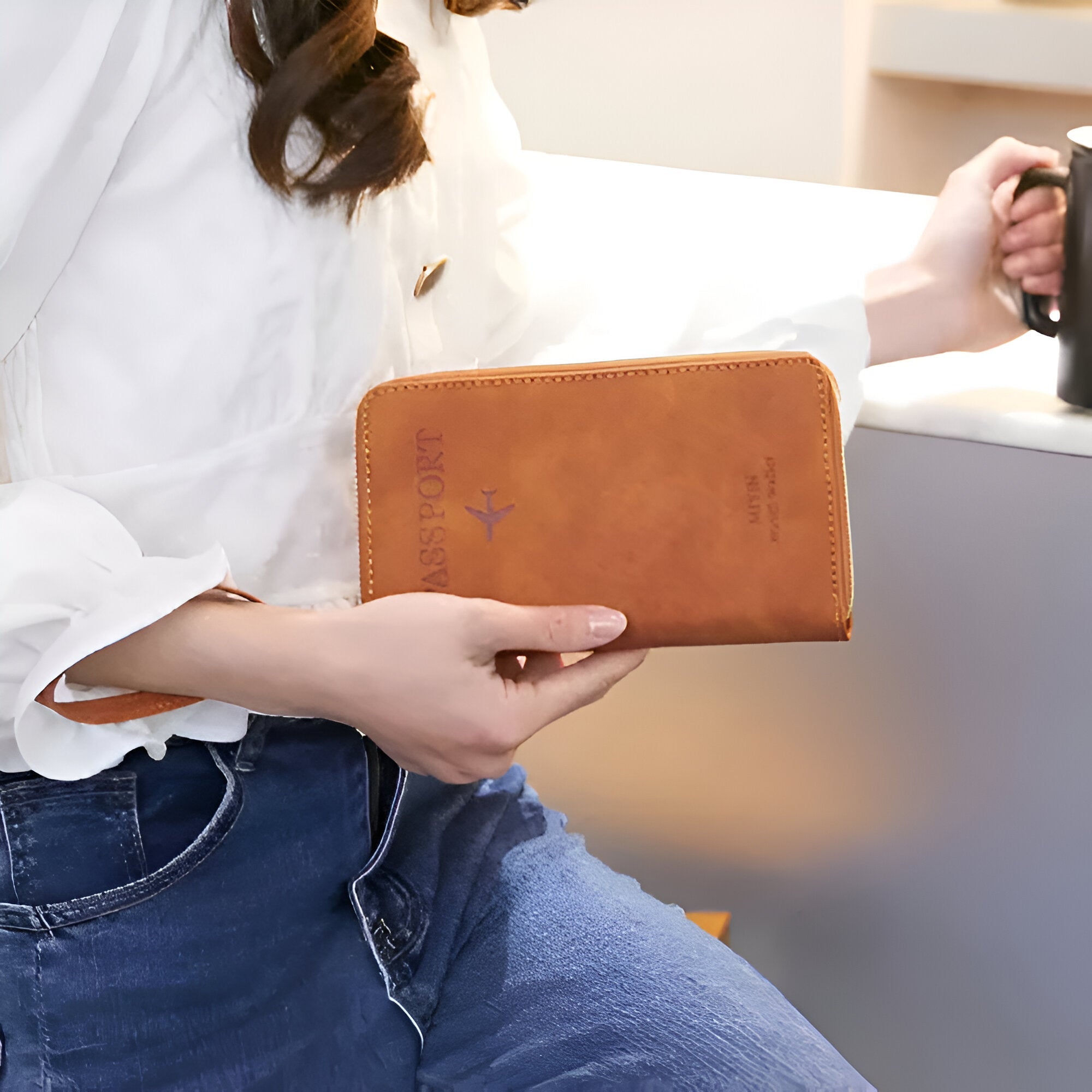 Woman holding a brown leather passport holder with embossed airplane design, wearing casual white blouse and jeans, sitting at a table with a coffee mug.