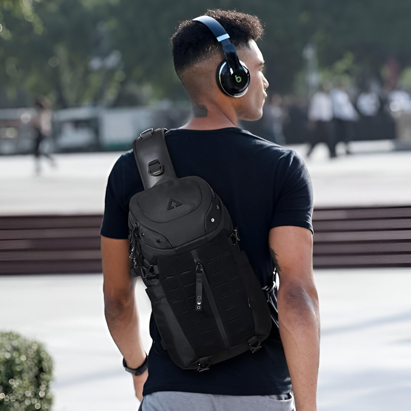 Man wearing black tactical backpack and headphones, walking outdoors in urban setting. Stylish, modern gear for travel and everyday use.