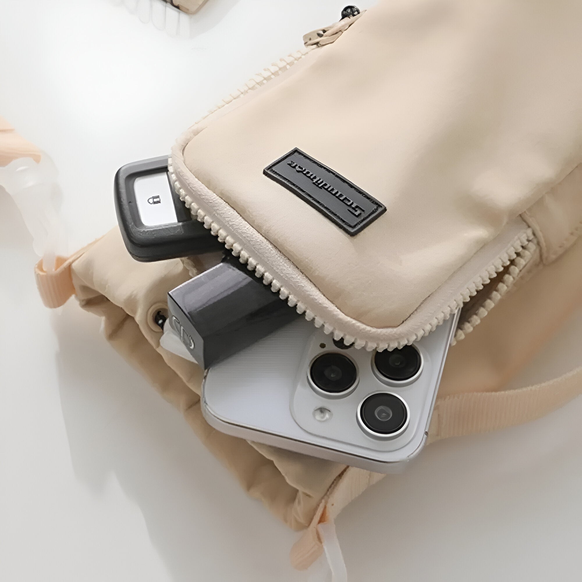 Beige crossbody bag with zipper, containing a smartphone, USB drive, and key fob. Perfect for tech-savvy travelers. Stylish and functional accessory.