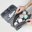 Hands organizing toiletries in a gray travel toiletry bag with multiple compartments, featuring bottles, a toothbrush, and a razor. Ideal for travel essentials.