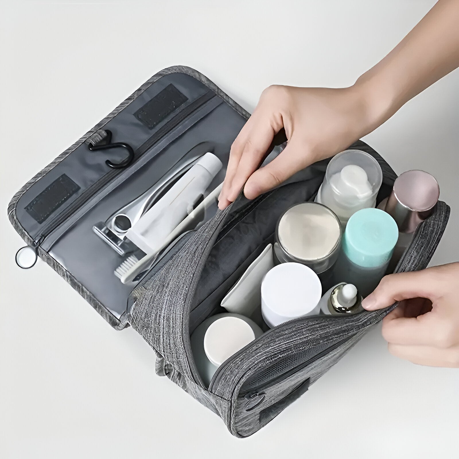 Hands organizing toiletries in a gray travel toiletry bag with multiple compartments, featuring bottles, a toothbrush, and a razor. Ideal for travel essentials.