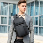 Man wearing a stylish gray suit and carrying a sleek black crossbody backpack, standing in front of modern glass building. Fashionable urban style.