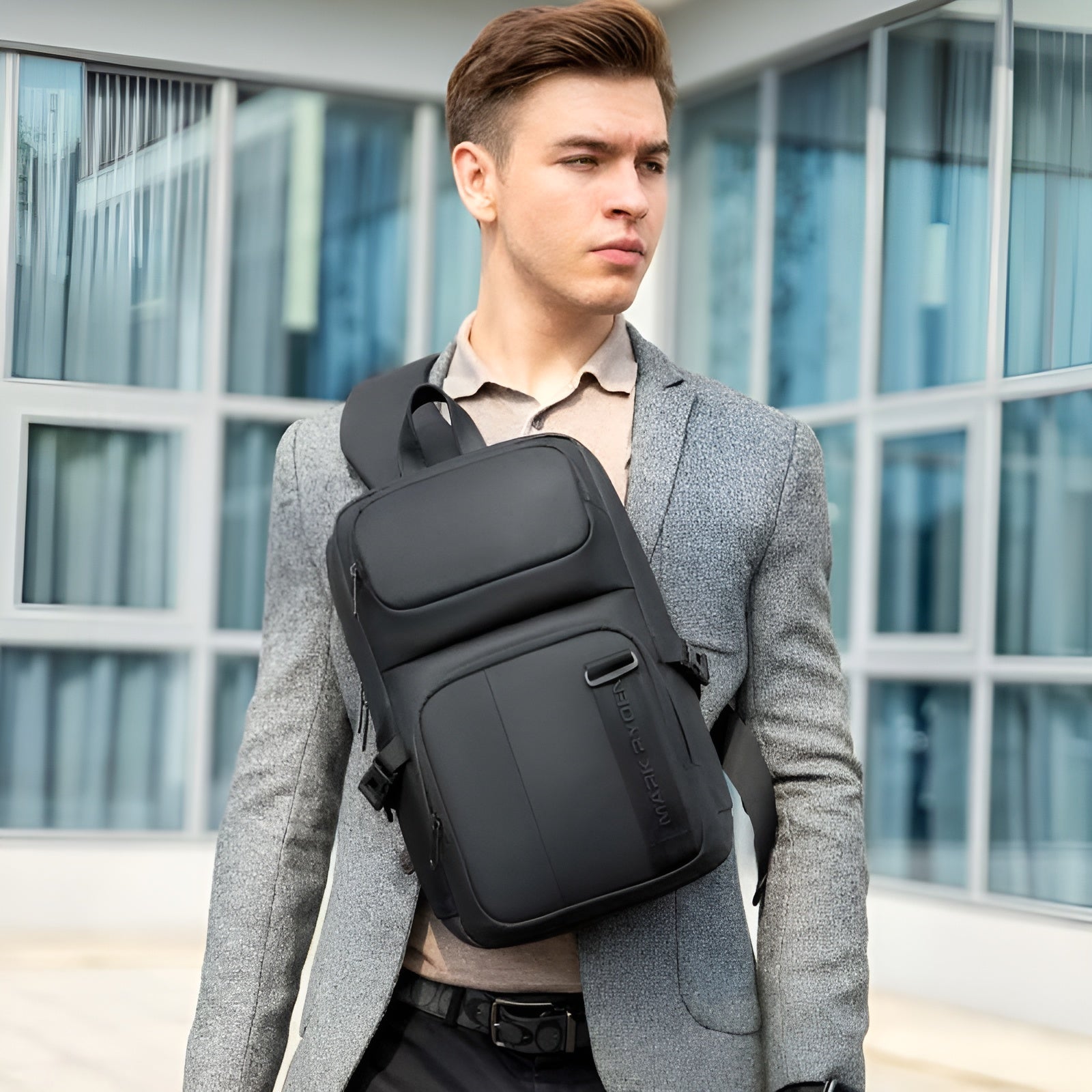 Man wearing a stylish gray suit and carrying a sleek black crossbody backpack, standing in front of modern glass building. Fashionable urban style.