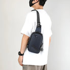 Man wearing a black crossbody sling bag with adjustable strap, paired with a white t-shirt and beige pants. Fashionable men's accessory.