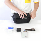 Person organizing items into a black travel pouch on a white surface, including a smartphone, watch, cardholder, and small case.