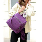 Woman holding a stylish purple backpack with multiple zippered compartments, wearing a casual white jacket and black pants. Fashionable urban accessory.