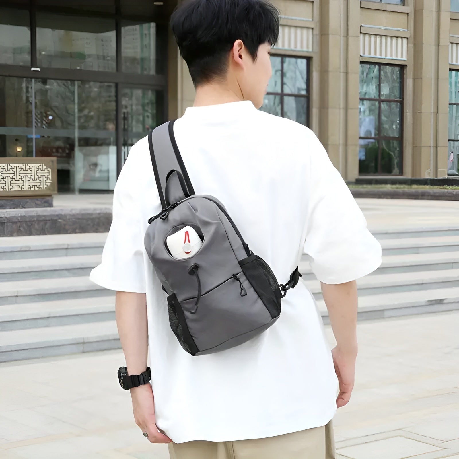 Man wearing a gray crossbody backpack with a white plush toy peeking out, standing outdoors in casual attire. Urban fashion, stylish accessory.