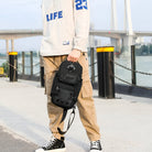 Person holding a black tactical sling bag, wearing a white hoodie and beige cargo pants, standing on a bridge walkway. Urban fashion, casual style.