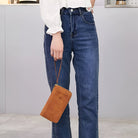 Woman holding a brown leather passport holder with wrist strap, wearing blue jeans and white blouse. Travel accessory, stylish passport wallet.