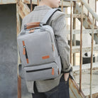 Stylish gray backpack with leather accents on a person in plaid shirt, standing on rustic stairs. Perfect for travel, school, or work essentials.