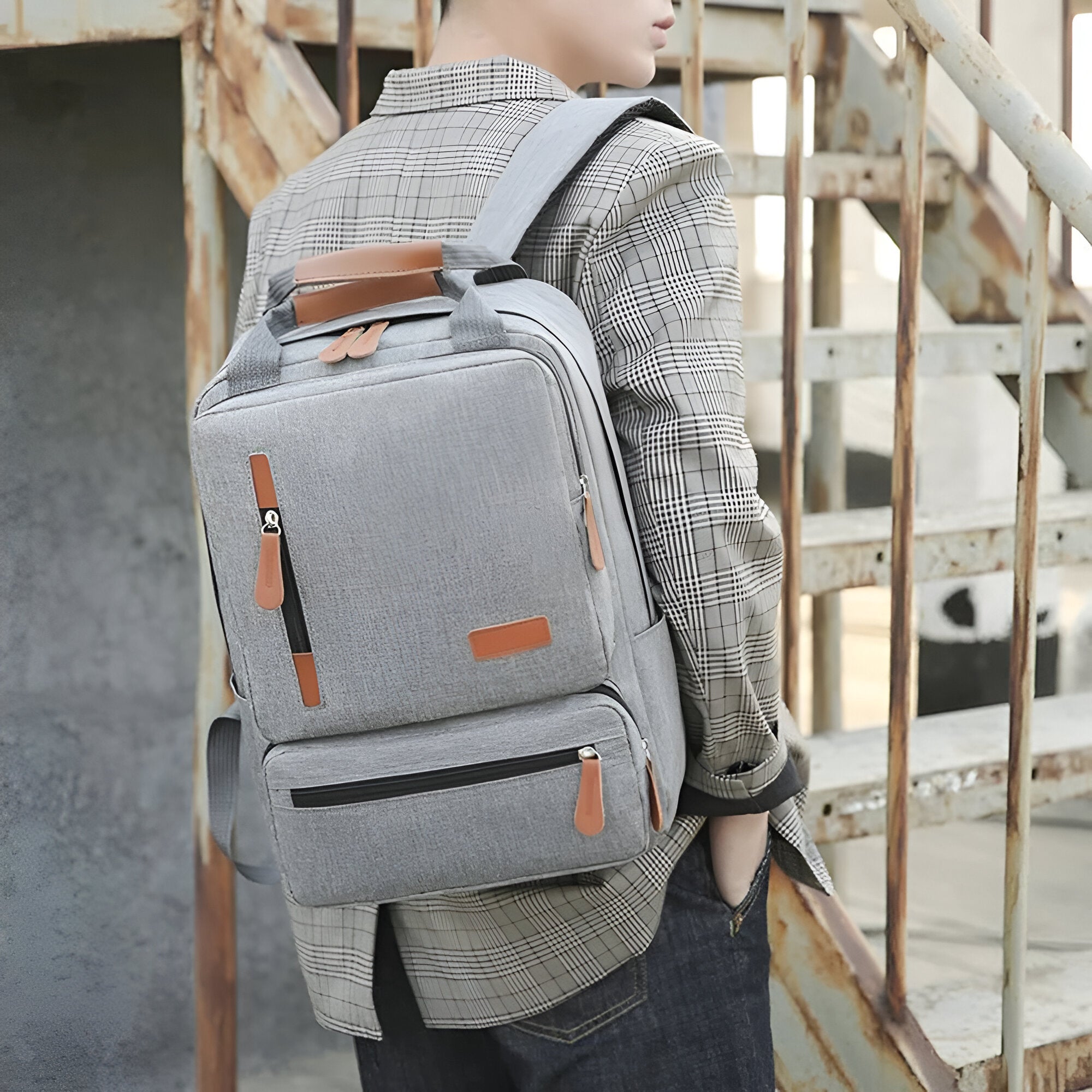 Stylish gray backpack with leather accents on a person in plaid shirt, standing on rustic stairs. Perfect for travel, school, or work essentials.