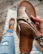 Beige leather sandal with braided strap held by a hand, worn with ripped jeans on a wooden floor and white rug. Fashionable women's footwear.