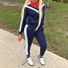 Woman wearing a navy blue classic tracksuit set with white and red accents, standing outdoors on a sidewalk. Fashionable athletic wear, casual style.