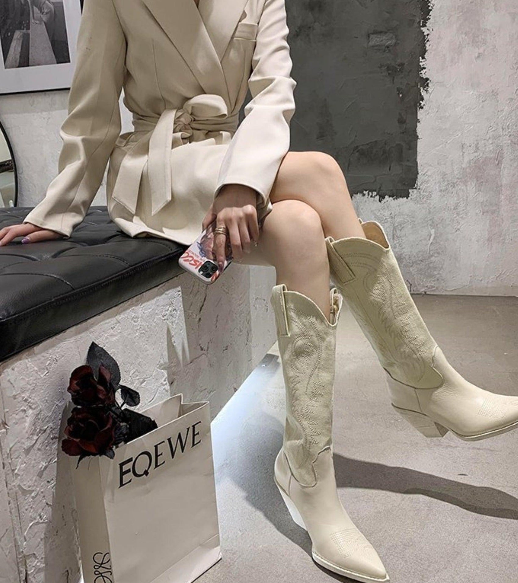 Knee-high white leather cowboy boots for women, featuring intricate stitching, paired with a stylish beige coat. Fashionable footwear for modern trends.