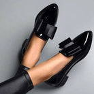 Shiny black patent leather loafers with large decorative bows, worn with black leggings, on a gray background. Fashionable women's footwear.