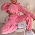 Woman in a pink cropped sweatshirt and joggers set, sitting on a clear chair, wearing white sneakers. Trendy loungewear fashion, casual style.