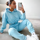 Woman in blue cropped sweatshirt and joggers, sitting on carpet, taking a mirror selfie. Casual loungewear, white sneakers, modern fashion.