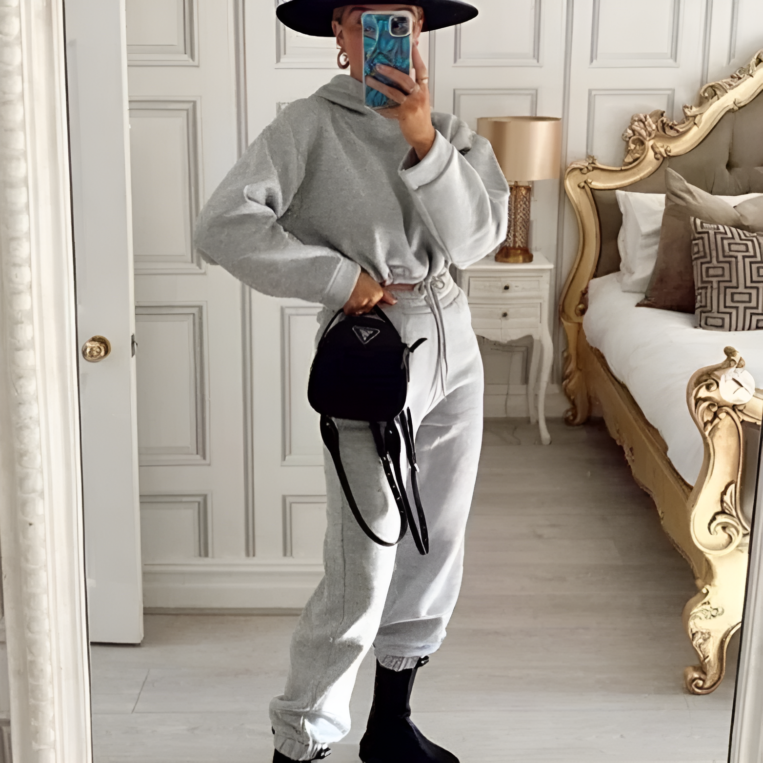 Woman in gray casual tracksuit and black boots, posing in elegant bedroom with ornate furniture, holding a black handbag, wearing a wide-brimmed hat.