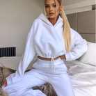 Woman in white cropped hoodie and sweatpants sitting on bed, casual loungewear fashion, cozy home outfit, stylish athleisure wear.