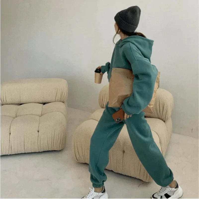 Woman in green sweatshirt set with beanie, holding coffee and bag, walking in modern living room. Cozy loungewear, casual fashion style.