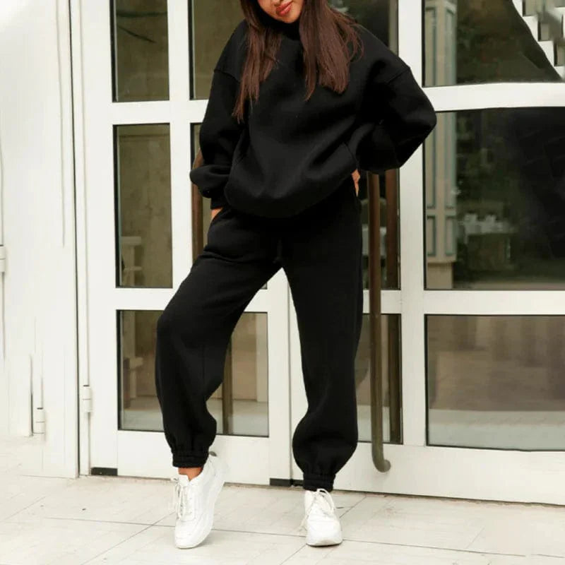 Woman wearing a black oversized sweatshirt and jogger set, standing in front of a glass door, paired with white sneakers. Casual fashion outfit.
