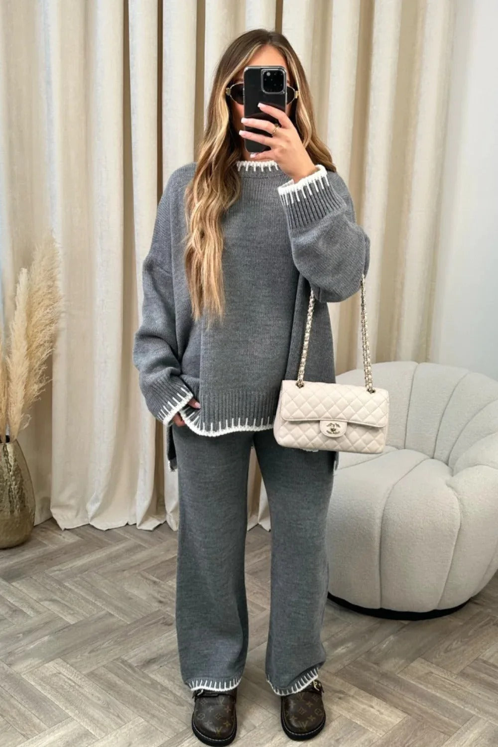 Woman in gray knit sweater and pants set, holding a white quilted handbag, taking a mirror selfie. Cozy fashion, casual outfit, stylish loungewear.