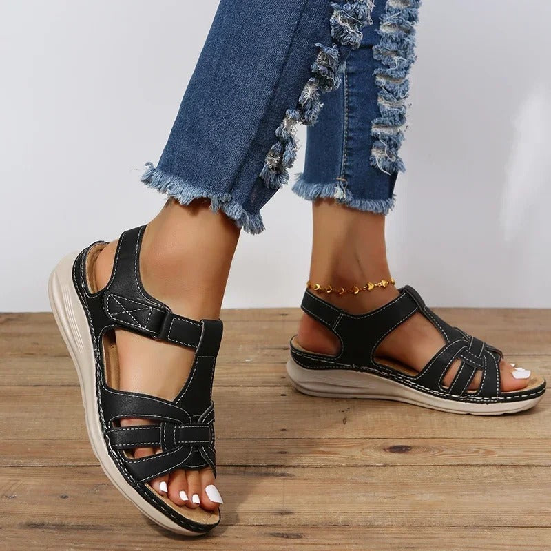 Black orthopedic sandals with adjustable straps on a wooden floor, worn with distressed denim jeans. Comfortable footwear for women.